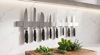 Nueve cuchillos de cocina con mangos negros en un soporte magnético metálico. Debajo, una tabla de cortar de madera con perejil, albahaca y tomillo.