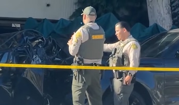 Dos agentes con chalecos tácticos permanecen junto a un automóvil negro destruido detrás de una cinta amarilla de seguridad