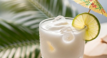 Primer plano de un vaso de agua de coco cremosa con hielo, una rodaja de limón y un paraguas de cóctel. Hojas de palma verdes al fondo desenfocadas.