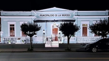 Edificio de la Municipalidad de La Merced de noche, con fachada celeste y reja de hierro forjado. Dos árboles y un coche oscuro se ven al frente