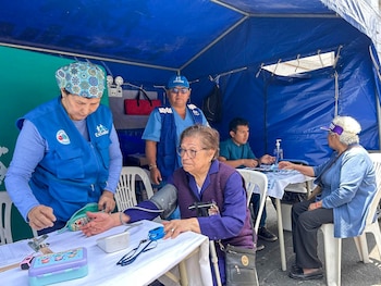 Una enfermera con chaleco azul mide la presión de una mujer sentada bajo una carpa médica azul junto a otros trabajadores y pacientes