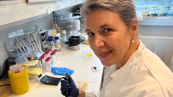 Mujer científica en bata de laboratorio y guantes azules, sonriendo a la cámara mientras sostiene una pipeta en un laboratorio con tubos de ensayo