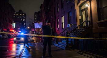 Oficial de policía en Nueva York muestra una bolsa con botellas marrones frente a un edificio residencial acordonado, con luces de patrullas encendidas de noche.