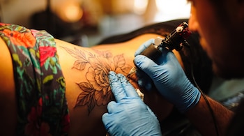 Una persona con guantes azules tatúa un diseño de flores en la espalda de otra persona. Una máquina de tatuar está en su mano.