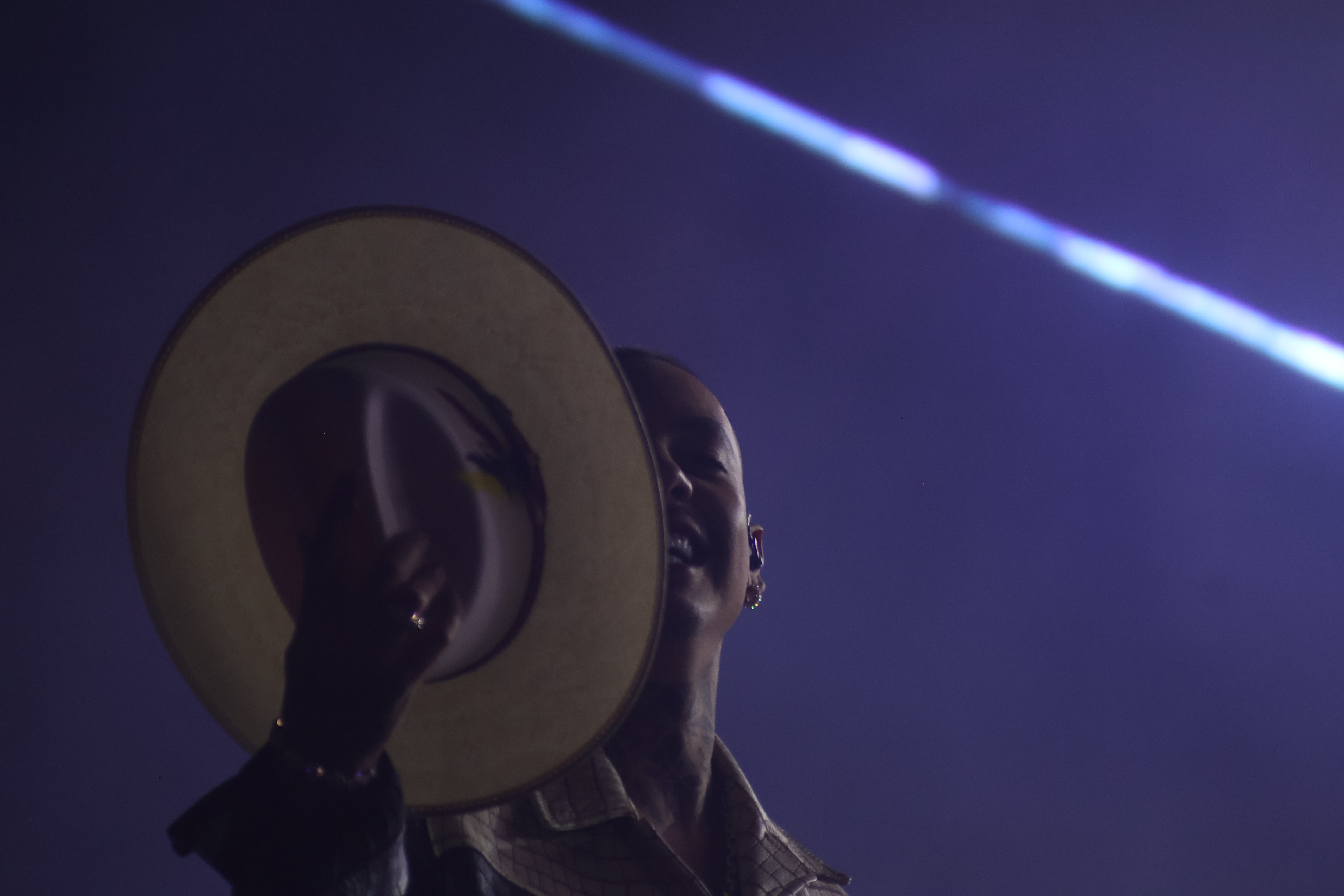 Un músico sostiene un sombrero frente a su rostro mientras sonríe en el escenario bajo una iluminación de tono violeta. (Selene Miranda/Infobae México)