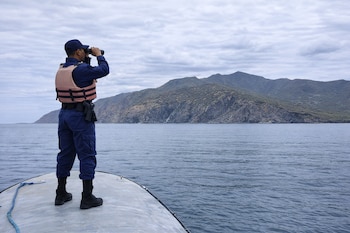 Hombre de espaldas con uniforme azul, gorra y chaleco salvavidas en un bote, usa binoculares mirando hacia montañas en el horizonte y el mar.