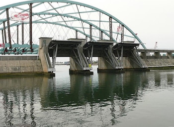 Un puente atirantado de color turquesa y gris, con una estructura de compuertas de metal y concreto debajo, sobre un cuerpo de agua. El cielo está nublado