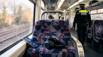 Asiento de tren con un teléfono móvil y auriculares negros en primer plano, y un agente policial desenfocado de espaldas en el pasillo al fondo.