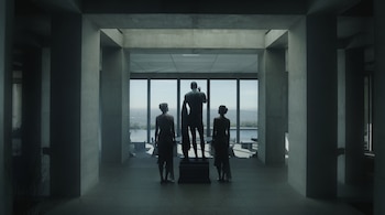 Dos personas y una estatua masculina vistas de espaldas en una sala con columnas de concreto frente a ventanales con vista a una ciudad.