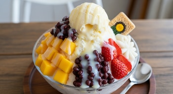 Un tazón de bingsu coreano en una mesa de madera, con hielo raspado, trozos de mango, fresas, frijoles rojos, helado y leche condensada, con un cracker decorativo.