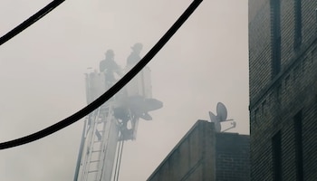 Dos bomberos en la cesta de una escalera extensible de camión, envueltos en humo denso. Un edificio de ladrillo se ve a la derecha y cables negros cruzan la imagen