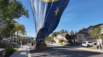 Globo aerostático azul y amarillo desinflándose en una calle residencial con casas y árboles. Varias personas asisten cerca de la cesta del globo