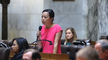 La Representante Francesca Hong, de ascendencia asiática, habla en un micrófono en un podio de madera en una sala de la asamblea con paredes de mármol y otras personas