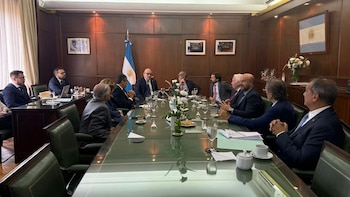 Un grupo de diez hombres sentados alrededor de una mesa de conferencias pulida. La sala tiene paneles de madera, cortinas y una bandera argentina