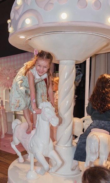 Niña ríe sobre caballo blanco de carrusel con silla rosa. Viste vestido floreado y tiene un tatuaje de Hello Kitty. Parte superior de carrusel y otras niñas