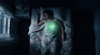 Vista cenital de una persona durmiendo en una cama a oscuras, cubierta hasta la cintura por una colcha. Un brillo verde emana de su pecho.