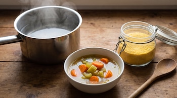 Olla de metal con agua humeante, tazón blanco con caldo de verdura, frasco de cristal con polenta amarilla, cuchara de madera sobre superficie de madera.