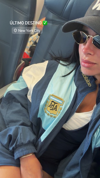 Una mujer sentada en un avión lleva una gorra oscura, gafas de sol y una chaqueta con el escudo de la Asociación del Fútbol Argentino (AFA). Se lee "New York City"