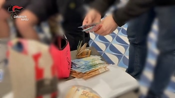 Primer plano de manos contando fajos de billetes de euro de 20 y 50, junto a una bolsa rosa y un logotipo de Carabinieri Nápoles en la esquina superior izquierda