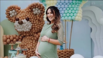Una mujer embarazada vestida de verde sonríe de pie frente a un oso hecho con globos marrones y una figura de globo aerostático