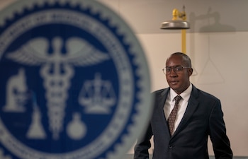 Un hombre con traje y gafas posa mirando al frente, con un sello azul redondo difuminado que incluye un caduceo en primer plano