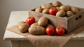 Caja de madera rústica con papas, con varios tomates rojos brillantes y más papas esparcidas en una mesa de madera clara con un trozo de yute.