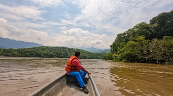 Un hombre con chaleco salvavidas naranja sentado en la proa de una canoa sobre un río turbio rodeado de vegetación y montañas.