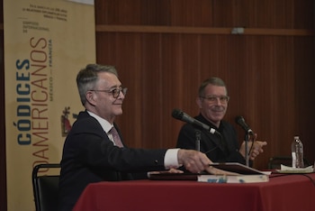 Dos hombres con gafas sentados ante una mesa con mantel rojo y micrófonos, junto a un panel informativo