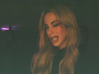 Primer plano de Tini Stoessel con cabello rubio largo, guiñando un ojo y sacando la lengua levemente, sobre un fondo oscuro con luz morada