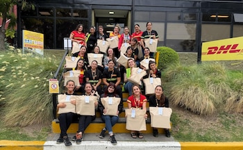 Un grupo de mujeres sonrientes del equipo DHL, algunas con uniforme, sentadas en escaleras, sostienen bolsas beige frente a un edificio con el logo de la empresa