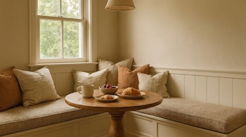 Rincón de desayuno acogedor con mesa redonda de madera. Incluye dos cruasanes, un cuenco de bayas, una taza y bancos acolchados junto a una ventana.