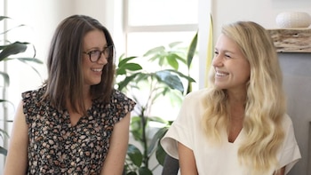 Dos mujeres, una con cabello castaño y gafas, y otra rubia, sonríen y se miran en un interior luminoso con plantas de fondo