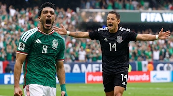 Dos jugadores de fútbol con camisetas verde y negra gritan de alegría en un campo. El de negro tiene los brazos abiertos. Fondo borroso de estadio.