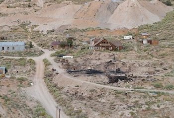 Camino de tierra entre casas de madera, un área quemada con escombros oscuros y colinas áridas con montículos de tierra