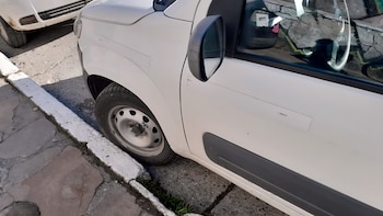 Vista lateral de un automóvil blanco estacionado con una rueda contra el cordón de una vereda. Se observa la calle de asfalto y el lateral de otro vehículo