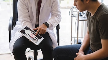 Un médico con bata blanca y corbata señala un diagrama del cuerpo humano con un área resaltada en rojo en una tablet, frente a un paciente atento.