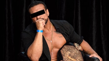 Hombre con barba, camisa negra abierta, rosario y cinturón de campeonato sentado en silla oscura. Lleva pulsera azul y tiene la mano en la boca