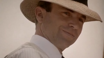 Primer plano de un hombre con sombrero de paja claro, camisa blanca y corbata oscura, que mira al frente con una leve sonrisa