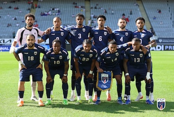Once futbolistas masculinos de Cabo Verde posan en el campo. Diez visten uniformes azul oscuro, uno de rosa pálido. Un jugador sostiene un banderín de la FCF
