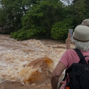 Una mujer de espaldas con sombrero y mochila toma una fotografía con un teléfono móvil hacia un río correntoso de agua marrón y vegetación