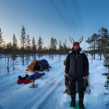 Hombre con chaqueta oscura, gorro rojo y astas de reno, pieles al hombro, parado en nieve con raquetas. Tienda de campaña y pinos al fondo, cielo azul