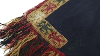 Primer plano de un textil Paracas con fondo oscuro, un borde beige adornado con figuras estilizadas rojas y verdes, y flecos multicolores