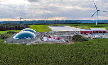 Vista aérea de dos biodigestores de cúpula verde, un edificio rojo, una laguna y campos agrícolas. Varias turbinas eólicas se alzan al fondo