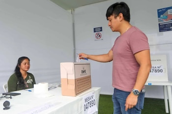 Hombre de camiseta rosa introduce papeleta en urna de votación con logo ONPE; mujer sentada de perfil en el fondo lo observa en mesa electoral