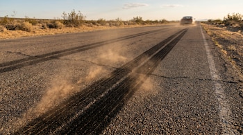Carretera asfaltada con profundas huellas de neumáticos y una nube de polvo. Un coche borroso se aleja en el horizonte de un paisaje desértico con arbustos.