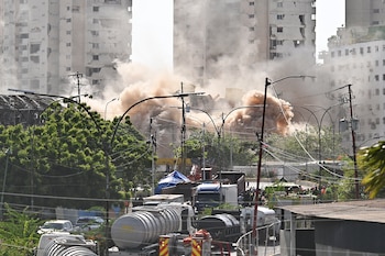 Edificios con daños visibles, una gran nube de polvo, árboles verdes, postes y cables eléctricos, varios camiones cisterna y personas en un área urbana