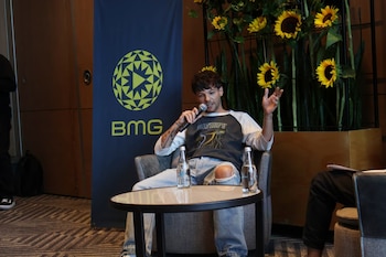 Louis Tomlinson, sentado en una silla, sostiene un micrófono y gesticula. Detrás, un estandarte de BMG y girasoles decorativos