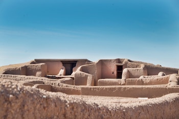 Conjunto de estructuras de adobe antiguas de tonos tierra con paredes, aberturas y entradas, bajo un cielo azul despejado, con detalles de ruinas en primer plano
