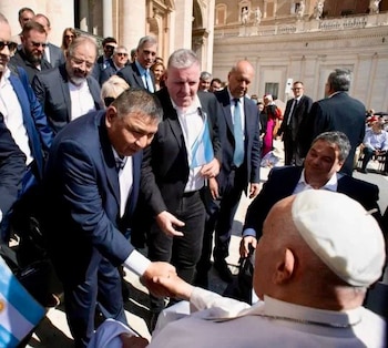 Jorge "Loma" Ávila, vestido con traje, se inclina para estrechar la mano del papa Francisco, quien está sentado en una silla de ruedas al aire libre, rodeado de otras personas