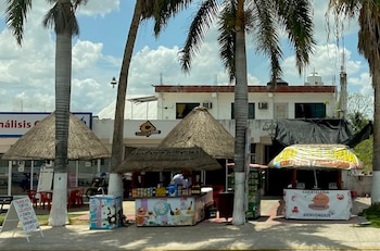 Tres palmeras, varios puestos de venta con techos de palma, edificios de fachada blanca, un letrero de café, un cartel de bebidas y alimentos, cielo con nubes